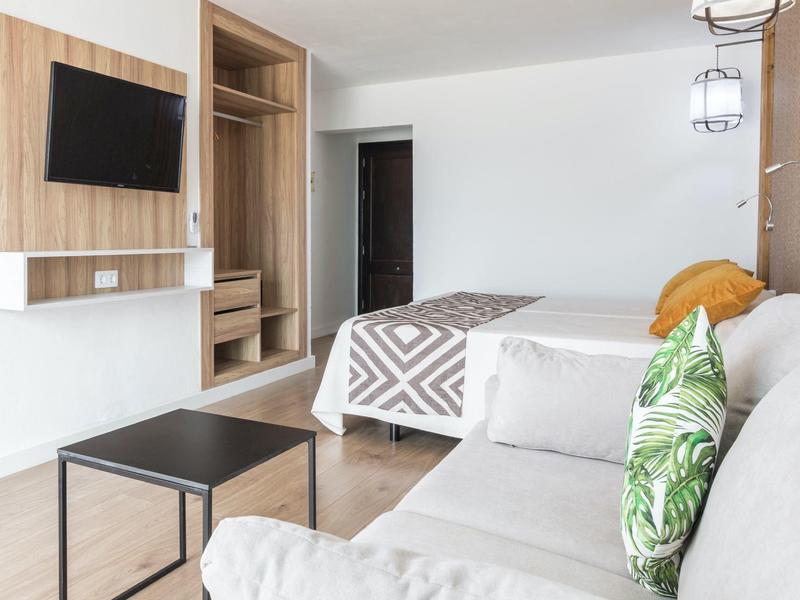 Helles Zimmer mit Bett, grauem Sofa, grünem Musterkissen, schwarzem Tisch und Wandfernseher.