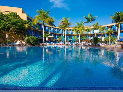Großer Pool mit blauer Umrandung umgeben von Palmen und Hotelgebäuden bei Sonnenschein.