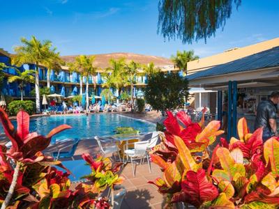 Poolbereich eines Hotels mit Sonnenschirmen, Palmen und bunten Pflanzen im Vordergrund.