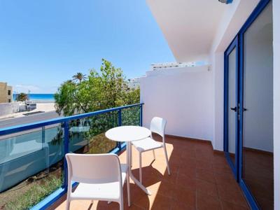 Balkon mit zwei weißen Stühlen und Tisch, Blick auf Meer und umliegende Gebäude bei klarem Himmel.