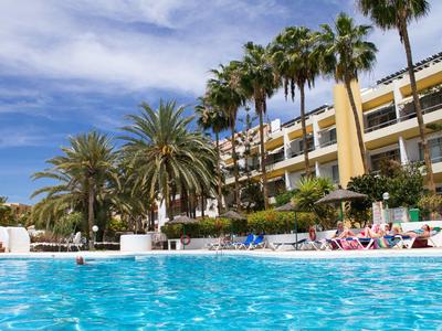 Hotel con grande piscina, lettini e palme sotto un cielo azzurro.
