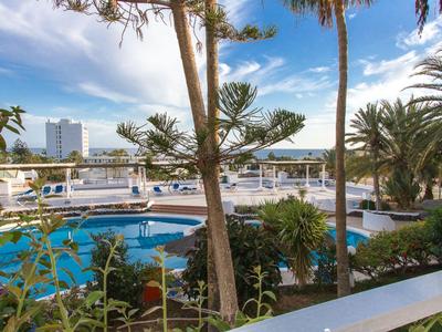Vista su un'area piscina con palme e hotel sullo sfondo in una giornata di sole.