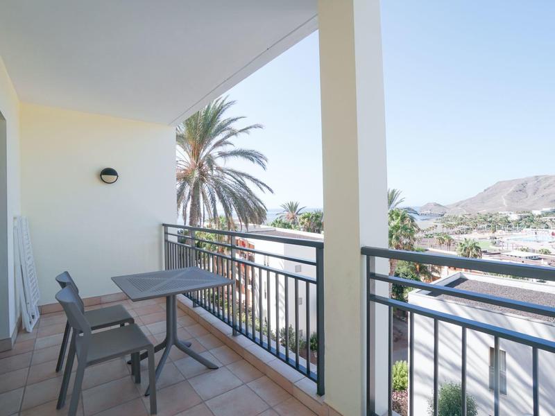 Balcone con due sedie e un tavolo, vista su palme e montagne sotto un cielo limpido.