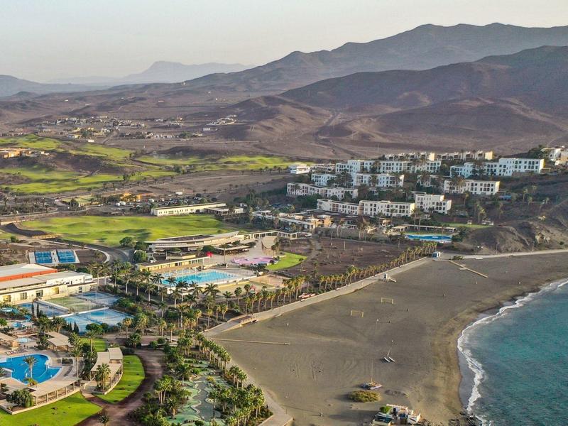 Vista aerea di una località costiera con piscine, hotel e paesaggio montano sullo sfondo.