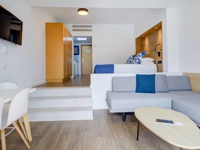 Modern hotelkamer met zithoek, tv, kitchenette en eethoek in een lichte stijl.