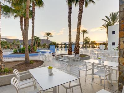 Area esterna dell'hotel con tavoli, sedie, piscina e palme al tramonto.