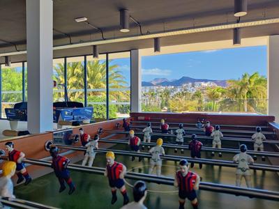 Een tafelvoetbal staat in een zonnige kamer met grote glazen ramen met uitzicht op palmbomen en bergen.