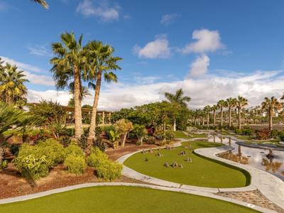 Park-like garden with palm trees, green lawns, and a water stream under blue sky.