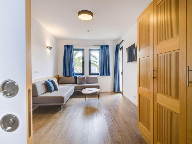 Uitzicht op een lichte hotelkamer met bank, tafel en grote ramen met blauwe gordijnen.