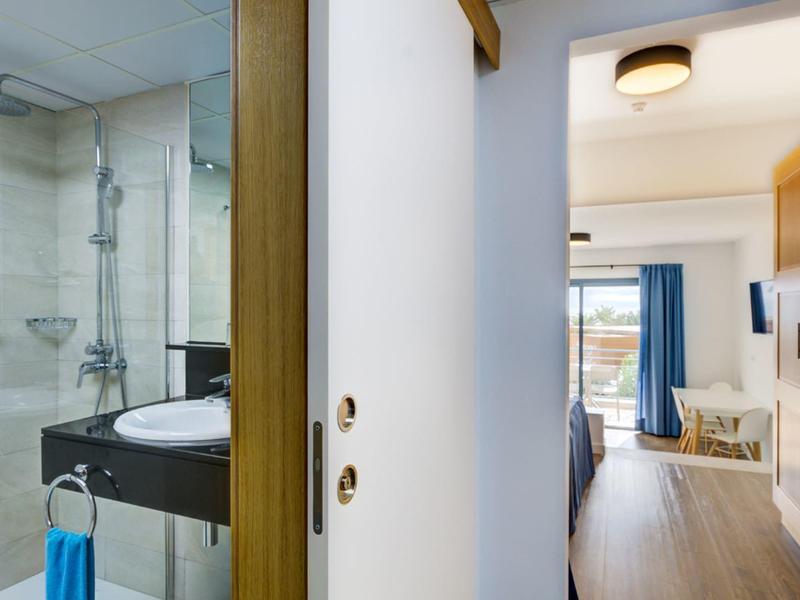 Uitzicht vanuit moderne badkamer met douche naar lichte hotelkamer met balkon en zithoek.