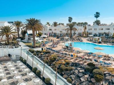 Complejo hotelero con piscina, sillas de descanso y edificios blancos bajo cielo azul claro.