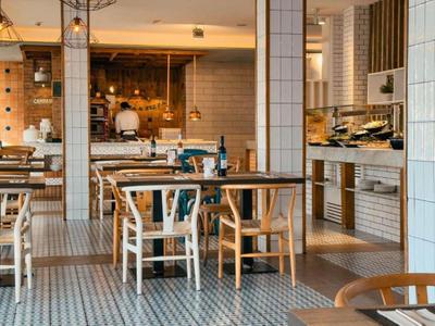 Área de comedor moderna en hotel con mesas de madera y suelo de baldosas en tonos neutros.
