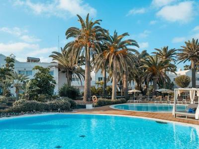 Gran piscina azul de hotel rodeada de palmeras y edificios blancos bajo un cielo azul.