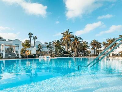 Gran piscina exterior con tobogán y palmeras frente a edificio de hotel claro bajo cielo azul.