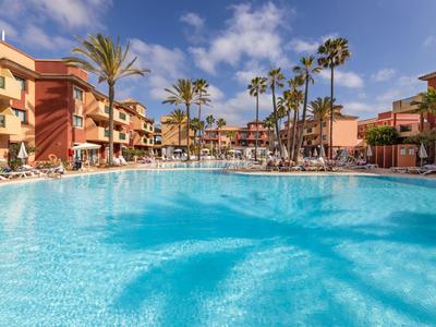 Gran piscina del hotel con palmeras y habitaciones soleadas bajo un cielo azul.