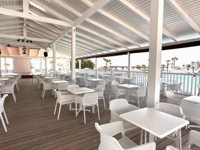 Světle zastřešená restaurace s bílými židlemi a stoly, výhled na bazén a palmy.