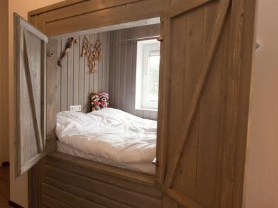 Holzstruktur mit offener Tür zeigt ein gemütliches Bett mit weißer Bettdecke und Fenster im Hintergrund.