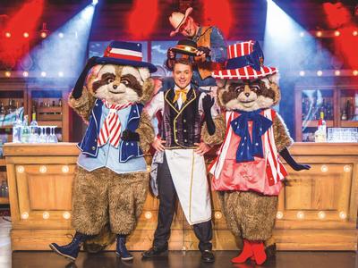 Zwei Maskottchen in patriotischen Kostümen und eine Person in klassischer Kellnerkleidung auf einer Bühne.