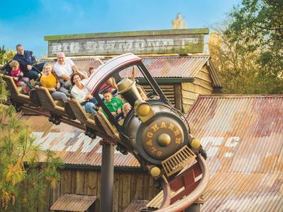 Menschen fahren in einer Vintage-Holzachterbahn in einem Freizeitpark mit einer alten Hütte im Hintergrund.
