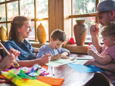 Familie sitzt am Holztisch, Kinder malen mit bunten Stiften, Mutter und Vater lachen.