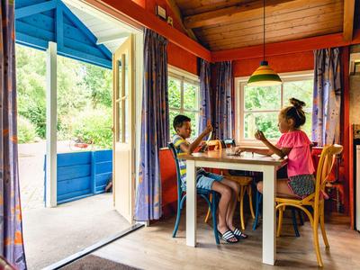 Zwei Kinder sitzen an einem Esstisch in einem bunten, gemütlichen Raum mit offener Tür zum Garten.