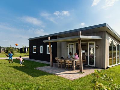 Modernes, dunkles Holzhaus mit Veranda und Garten, zwei Erwachsene am Tisch, Kinder spielen auf Gras.