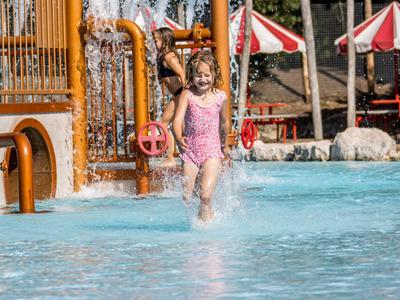 Ein Mädchen im rosa Badeanzug spielt im flachen Pool mit Wasserspielen und roten Schirmen im Hintergrund.