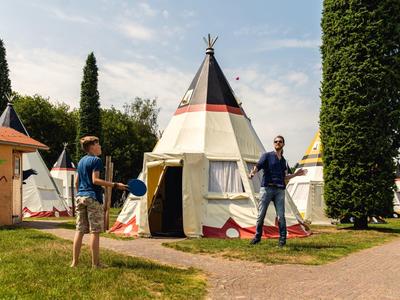Zwei Männer stehen vor weißen Tipis mit roten Mustern in einem sonnigen Park mit grünen Bäumen.