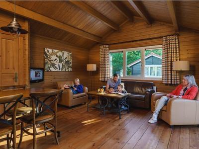 Gemütliches Wohnzimmer mit Holzwänden, großen Fenstern und drei Personen auf Sofas und Sesseln.