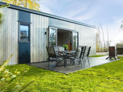 Modernes Holzhaus mit großer Glasschiebetür, Terrasse mit Tisch und Stühlen, umgeben von grünem Rasen.