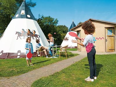 Drei Kinder spielen in einem Garten mit bemalten Tipis und einem Holzhaus im Hintergrund.