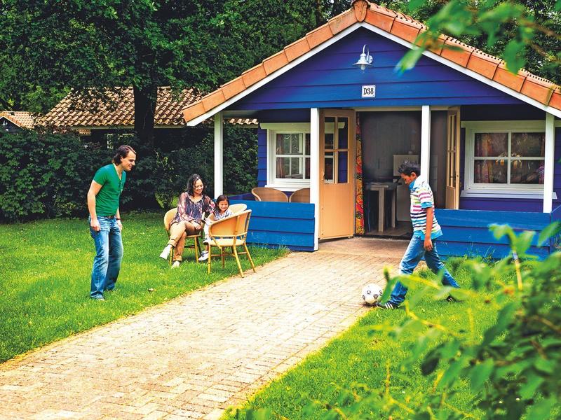 Familie vor blauem Holzhaus mit Garten, Rasen und gepflastertem Weg, grüne Bäume im Hintergrund.