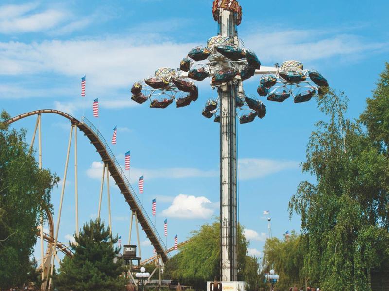 Freizeitpark mit Karussell, das in der Luft schwingt, umgeben von Gras, Bäumen und blauen Himmel.