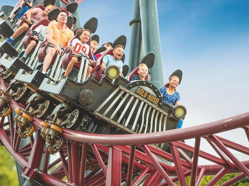 Mehrere Personen fahren lachend und aufgeregt eine rote Achterbahn bei klarem Himmel.