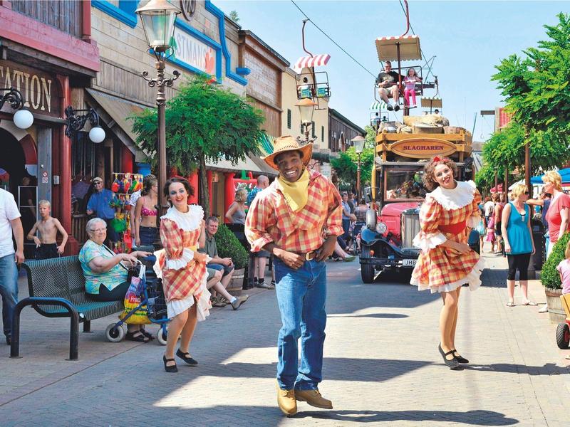 Straße mit tanzenden Menschen in bunter Kleidung, Sitzbänken und Geschäften bei sonnigem Wetter.
