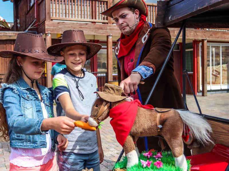 Kinder in Cowboy-Kleidung füttern ein Pony in einer Westernkulisse bei Sonnenschein.