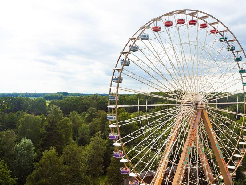 Ein großes Riesenrad mit bunten Gondeln steht neben grünen Bäumen unter einem bewölkten Himmel.