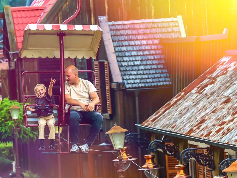 Mann und Kind sitzen auf einer Holzschaukel mit Hausdach und Sonnenlicht im Hintergrund.