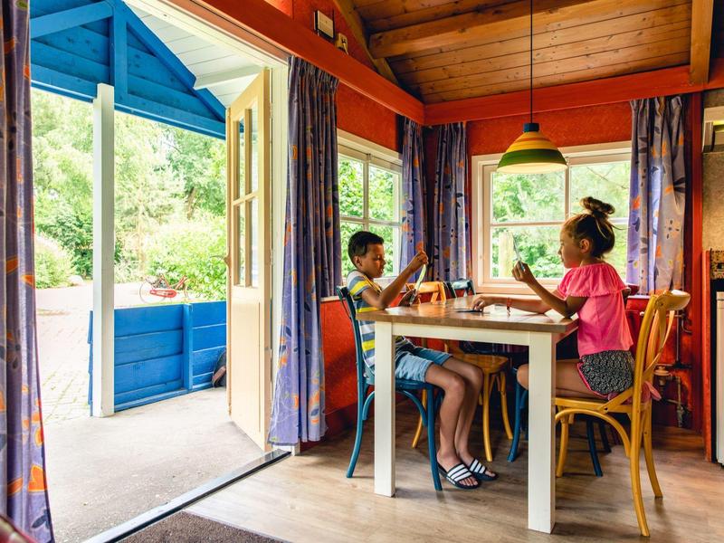 Zwei Kinder sitzen an einem Esstisch in einem bunten, gemütlichen Raum mit offener Tür zum Garten.
