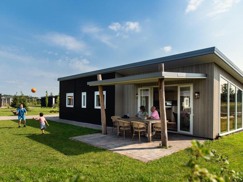 Modernes, dunkles Holzhaus mit Veranda und Garten, zwei Erwachsene am Tisch, Kinder spielen auf Gras.