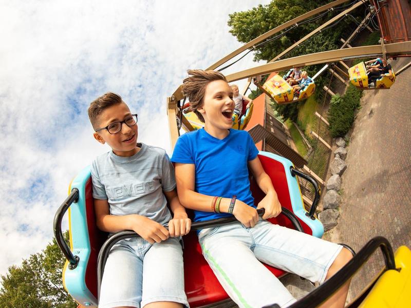 Zwei Kinder sitzen lachend in einer bunten Achterbahn mit Himmel und Bäumen im Hintergrund.