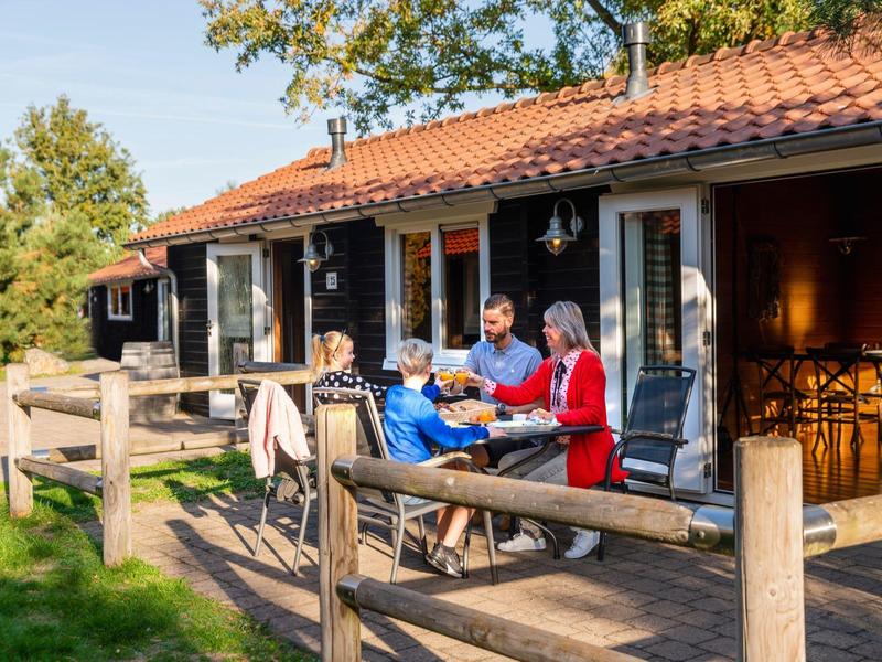 Drei Personen sitzen auf der Terrasse eines Holzhauses, umgeben von Grün und Holzgeländer.