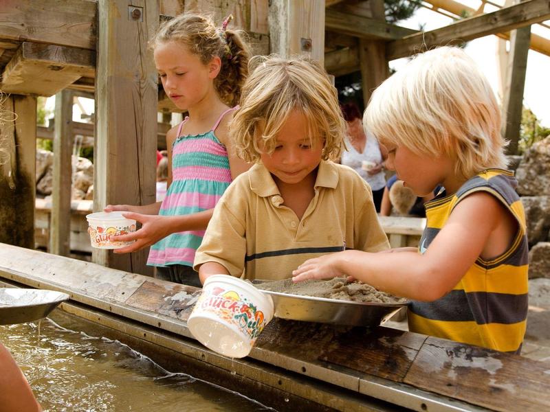 Kinder spielen mit Wasser an einem Holzgestell, zwei schöpfen Wasser mit Eimern und einem Sieb.