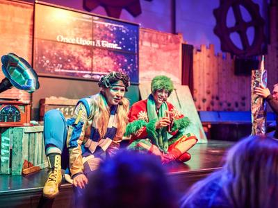 Two actors in colorful costumes sit on a stage with props.
