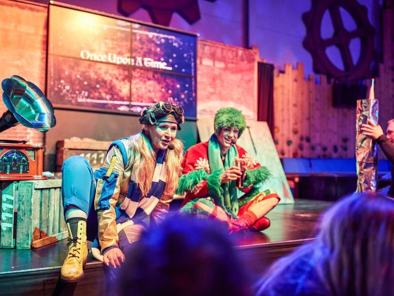 Two actors in colorful costumes sit on a stage with props.