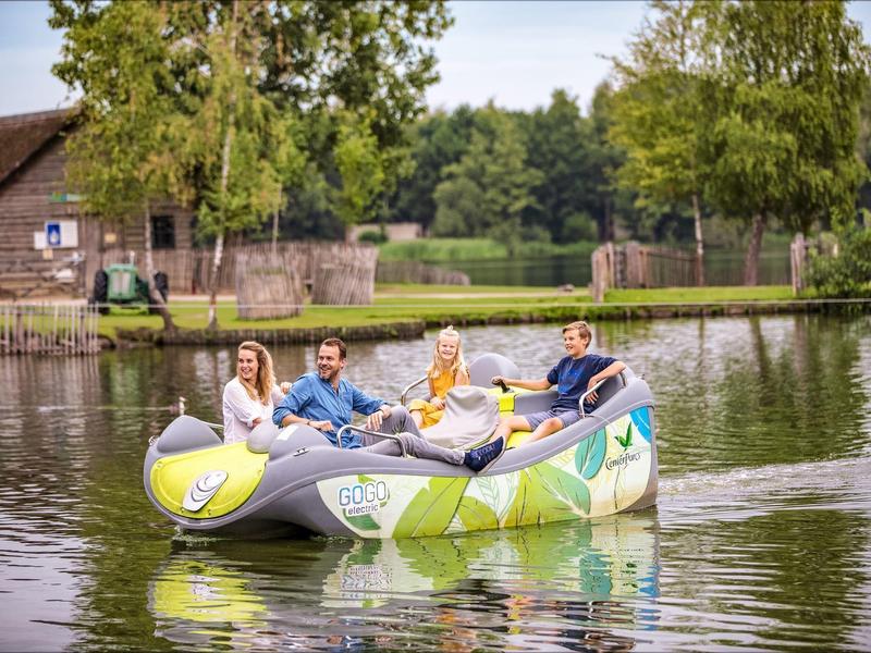 Four people sit in a colorful inflatable boat on a calm lake near a farm.