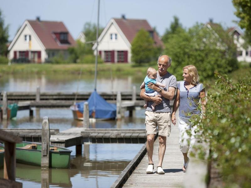 Oudere stel loopt met kleinkind op een steiger bij rustig water, huizen op de achtergrond zichtbaar.