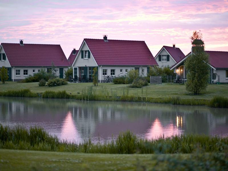 Meerdere beige vakantiewoningen met rode daken bij een vijver bij zonsondergang.