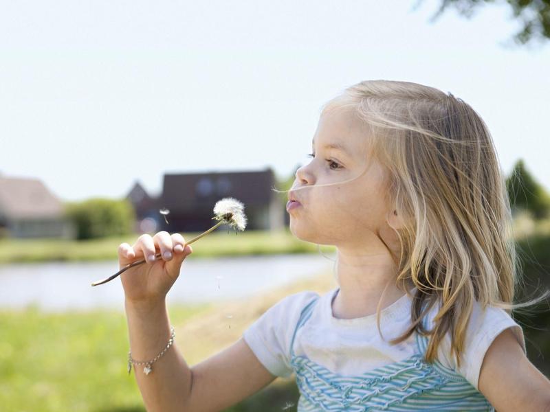 Meisje blaast op een paardebloem bij een meer met groen landschap.