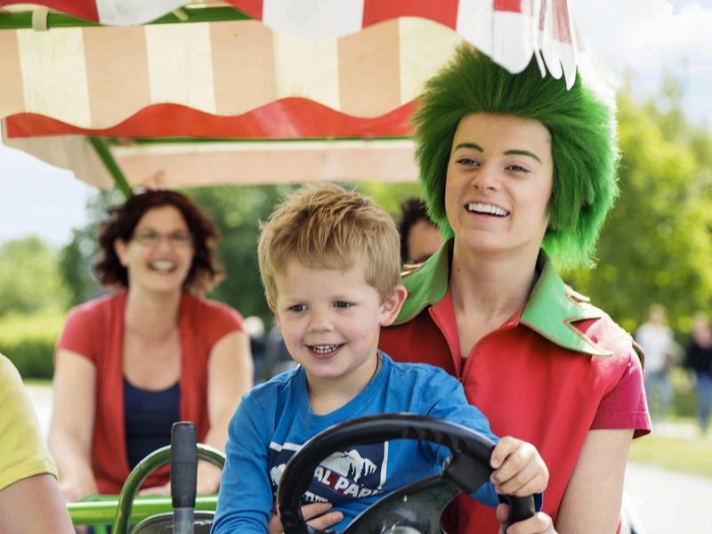 Blij gezin met kind dat rijdt op een kart in een pretpark onder een rood-witte zonwering.